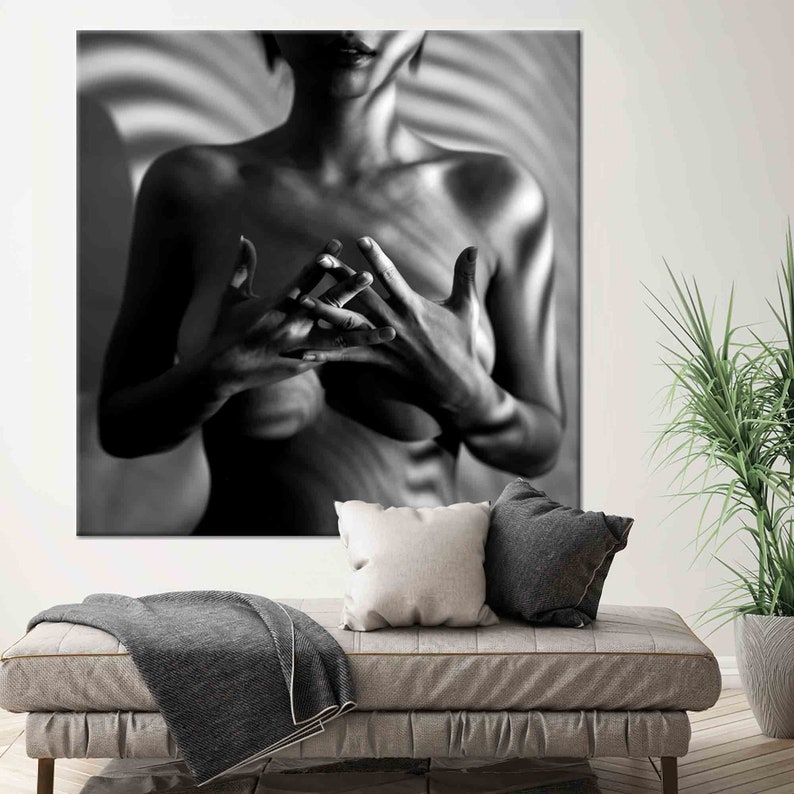 May include: Black and white art print of a person with hands clasped in front of the chest. The image is a close-up, with the person's upper body visible. The background has a striped pattern. The art print is displayed above a sofa with pillows and a blanket.