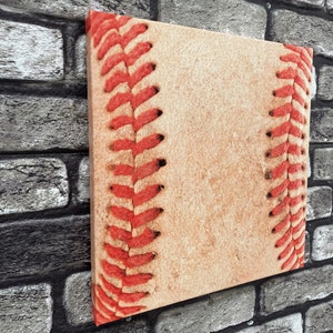 May include: Square canvas art featuring a close-up of a baseball. The artwork showcases the textured, light tan surface of the ball with vibrant red stitching along the seams. The piece is mounted on a wooden frame.
