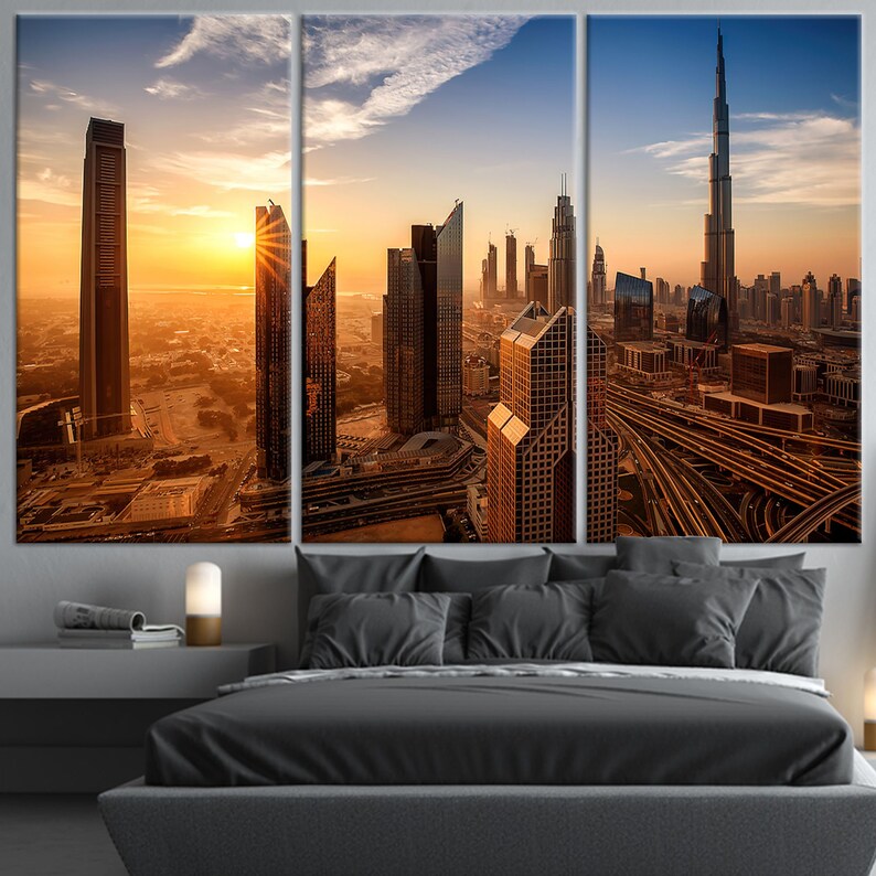 May include: A three-panel canvas print showing a cityscape at sunset. The image features modern skyscrapers, including the Burj Khalifa, bathed in a warm, golden light. The artwork is displayed above a bed with grey bedding.