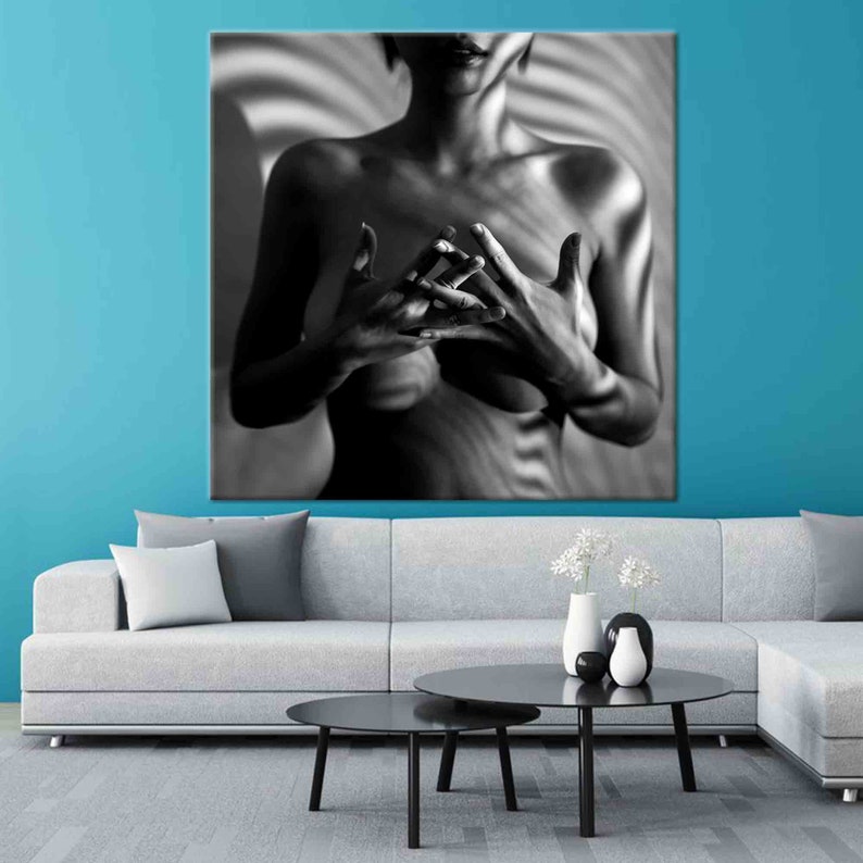 May include: Black and white canvas art print featuring a person with hands clasped in front of their chest. The background has a striped pattern. The artwork is displayed above a gray sofa and black coffee tables.