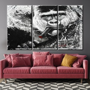 May include: A triptych canvas art piece featuring a black and white gorilla portrait with a cigar. The artwork has a splattered paint effect, with red accents on the cigar. The gorilla's face is detailed, with a contemplative expression. The art hangs above a pink sofa.