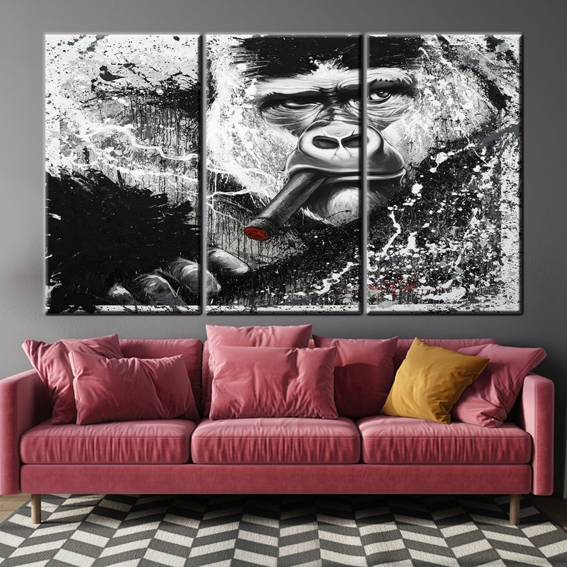 May include: A triptych canvas art piece featuring a black and white gorilla portrait with a cigar. The artwork has a splattered paint effect, with red accents on the cigar. The gorilla's face is detailed, with a contemplative expression. The art hangs above a pink sofa.