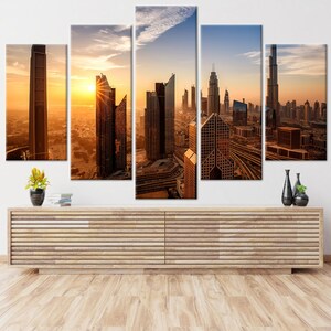 May include: A five-panel canvas print of a cityscape at sunset. The image shows modern skyscrapers in shades of brown and gold, with the sun setting in the background. The artwork is displayed above a wooden cabinet.