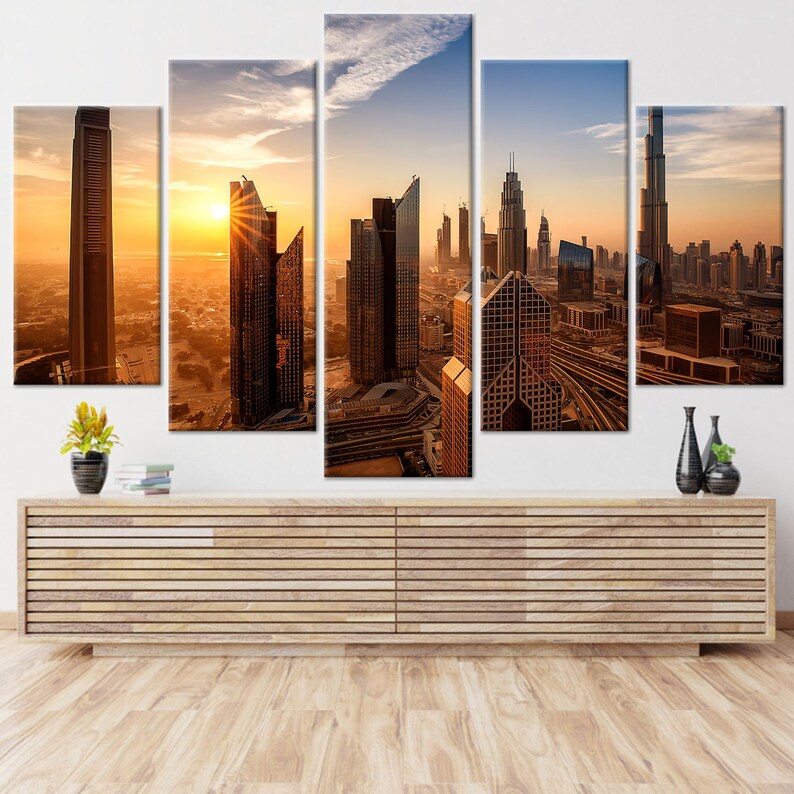 May include: A five-panel canvas print of a cityscape at sunset. The image shows modern skyscrapers in shades of brown and gold, with the sun setting in the background. The artwork is displayed above a wooden cabinet.