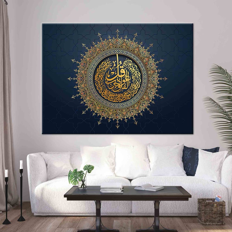 Puede incluir: Gran lienzo art&iacute;stico con caligraf&iacute;a &aacute;rabe dorada en un dise&ntilde;o circular ornamentado. La obra de arte est&aacute; sobre un fondo azul oscuro con un patr&oacute;n geom&eacute;trico. Se exhibe sobre un sof&aacute; blanco en un sal&oacute;n.