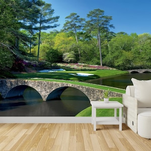 May include: A stone bridge over a pond with a golf course in the background. The bridge is surrounded by lush green trees and bushes. The pond is calm and reflective, and the sky is blue with white clouds.