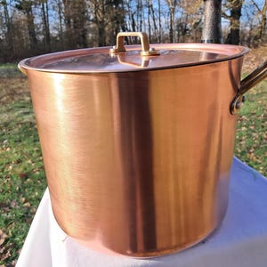 Puede incluir: Una olla grande de cobre con tapa y dos asas laterales. La olla tiene una superficie brillante y reflectante y un asa rectangular en la tapa. La olla está sobre una superficie blanca, con árboles al fondo.