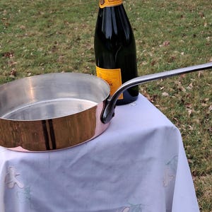 May include: A copper-bottomed, stainless steel frying pan with a long, silver handle. The pan is oval-shaped and sits on a white tablecloth. A dark bottle of champagne is in the background.
