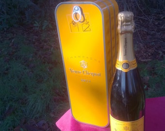 May include: A yellow gift box with a silver lid and a bottle of Veuve Clicquot Brut Champagne. The box has the Veuve Clicquot logo and the words "Champagne Veuve Clicquot Brut" printed on it.