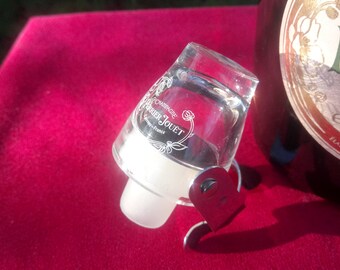May include: A clear glass champagne bottle stopper with a heart-shaped metal accent. The stopper has the words "Champagne Perrier Jouët" printed on it. The stopper is resting on a red surface next to a champagne bottle.