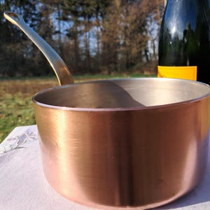 Può includere: Una casseruola di rame con un lungo manico color oro. L'interno è color argento. Sullo sfondo c'è una bottiglia scura con un'etichetta gialla. La casseruola è su una tovaglia bianca all'aperto.