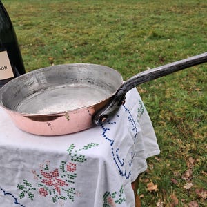 Puede incluir: Una sartén de cobre con mango negro. La sartén está sobre un mantel blanco con un patrón floral.