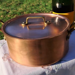 Puede incluir: Una olla de cobre con tapa y asas de latón. La olla es redonda y tiene una superficie brillante y reflectante. La tapa tiene un asa de latón. Una botella está en el fondo.