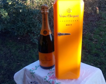 May include: A yellow Veuve Clicquot champagne bottle cooler with a black handle and the text "Veuve Clicquot Champagne Brut" on the front. A bottle of Veuve Clicquot champagne is next to the cooler.