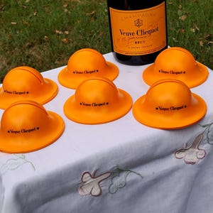 May include: Six orange, miniature hard hat-shaped coasters with the text "Veuve Clicquot" printed on them. A bottle of Veuve Clicquot champagne sits behind the coasters. The coasters are arranged on a white tablecloth with floral embroidery.