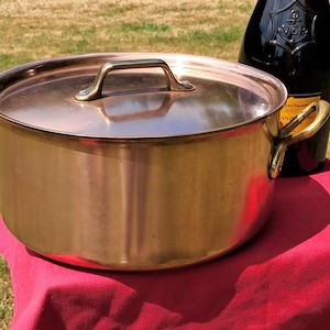 Puede incluir: Una olla de cocina de color cobre con tapa y asas de latón. La olla está sobre una superficie roja con una botella oscura en el fondo. La olla es redonda y tiene un acabado brillante. El diámetro de la olla es de unos 30 cm.