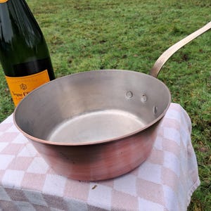 May include: A copper saucepan with a long, curved brass handle. The saucepan is empty and has a shiny, metallic finish.