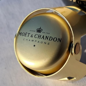 May include: A gold-colored champagne bottle stopper with the words "MOËT & CHANDON CHAMPAGNE" printed on the top. The stopper is made of metal and has a hinged design.