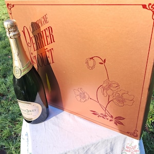 Könnte beinhalten: Eine Flasche Perrier-Jouët Champagner mit goldenem Etikett und einer großen, kupferfarbenen Schachtel. Die Schachtel zeigt das Perrier-Jouët-Logo und florale Illustrationen in Rot. Die Flasche ist dunkelgrün mit goldenem Hals.