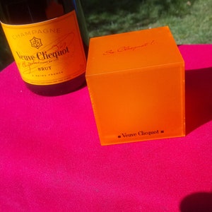 以下が含まれることがあります： ピンク色の表面に、ヴーヴ・クリコ・ブリュットのシャンパンボトルと、ヴーヴ・クリコ・ロゴが入ったオレンジ色の正方形の箱。シャンパンボトルには黄色のラベルが付いています。箱はオレンジ色です。