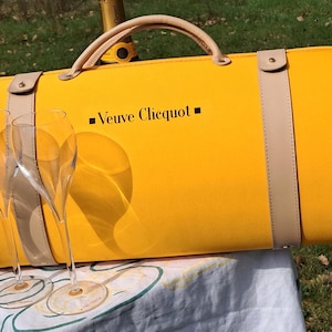 May include: A bright yellow Veuve Clicquot picnic bag with a tan handle and straps. The bag features the brand name. Two champagne flutes are in front of the bag.