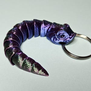 May include: A 3D-printed keychain shaped like a stylised sea serpent. The serpent is mainly purple with iridescent blue and green highlights. It has a silver key ring attached to its head.