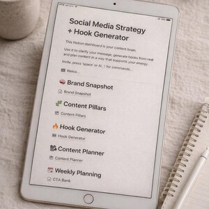 May include: A white tablet displays a "Social Media Strategy + Hook Generator" dashboard. The screen shows various content planning tools, including "Brand Snapshot," "Content Pillars," and "Hook Generator." A notepad and pen are next to the tablet.