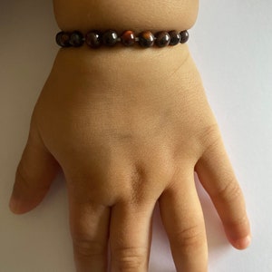 Peut inclure: Un bracelet de perles marron foncé et brun rougeâtre est porté au poignet. Le bracelet est composé de petites perles rondes. Le poignet repose sur une surface blanche. Les perles sont probablement en pierre naturelle.