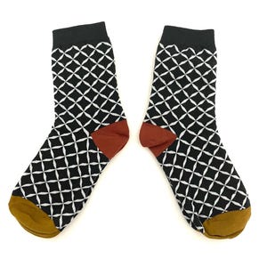 May include: A pair of crew socks featuring a black and white geometric diamond pattern. The socks have solid black cuffs, rust-colored heels, and mustard yellow toes. The design consists of repeating white four-pointed star shapes set against a black background.