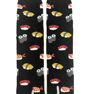 May include: A pair of black split-toe socks featuring a repeating pattern of various sushi pieces, including nigiri with red fish, yellow tamago, and grey maki rolls with green accents. The socks have a distinct separation between the big toe and other toes.