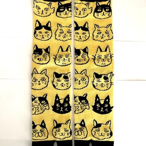 May include: A pair of yellow split-toe socks featuring a repeating pattern of black and white cat faces. The socks have a solid black cuff at the top and a solid black split-toe design at the bottom, with various feline expressions printed throughout.