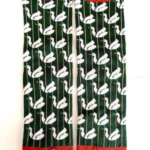 May include: A pair of split-toe socks featuring a dark green background with a repeating white swan pattern and thin vertical gold stripes. The socks have a solid red cuff at the top and a solid red split-toe section at the bottom.