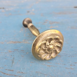 May include: A brass curtain tieback hook featuring a circular face with a raised, embossed floral petal design. The hardware includes a slender, tapered stem extending from the back of the decorative round plate, finished in a muted, aged metallic tone.