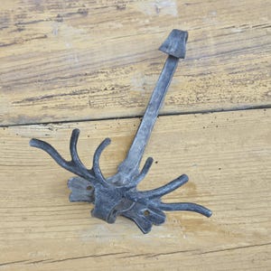 Puede incluir: Un gancho de pared de metal gris oscuro forjado a mano que presenta un brazo vertical central con un gancho superior y una sección inferior con forma de cornamenta ramificada. La base incluye dos orificios de montaje circulares.
