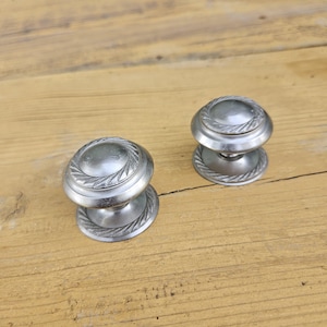 Può includere: Due pomelli rotondi per armadietti in metallo color argento spazzolato. Ogni pomello presenta un centro liscio e bombato circondato da un bordo decorativo testurizzato a corda lungo il bordo della faccia circolare e della base.