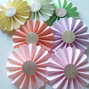 Puede incluir: Primer plano de una colección de flores de papel hechas a mano en varios colores pastel, incluyendo amarillo, verde, melocotón, rosa y morado. Cada flor tiene un centro plateado brillante.