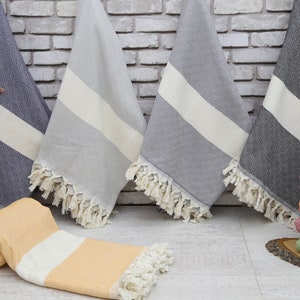 May include: Five Turkish towels with fringe, in various colors, including gray, blue, and orange, are displayed on a white brick wall background. The towels are folded and draped over the wall, with a stack of towels in the lower right corner.