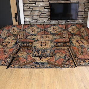 Może przedstawiać: Duża, bogato zdobiona sofa modułowa z pasującą otomaną i poduszkami do rzucania. Tapicerka ma szczegółowy, tradycyjny wzór w odcieniach czerwieni, niebieskiego i złota. Sofa jest ustawiona w kształcie litery U, tworząc przytulną część wypoczynkową.