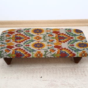 Puede incluir: Un reposapiés rectangular con un estampado floral en naranja, amarillo, azul y verde sobre un fondo claro. El taburete tiene patas de madera marrón oscuro. Una pequeña planta en maceta está a la derecha.