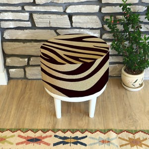 Puede incluir: Un pequeño taburete redondo con una parte superior con estampado de cebra en beige y marrón oscuro. El taburete tiene tres patas blancas y está colocado sobre un suelo de madera. A la derecha del taburete hay una pequeña planta en maceta.