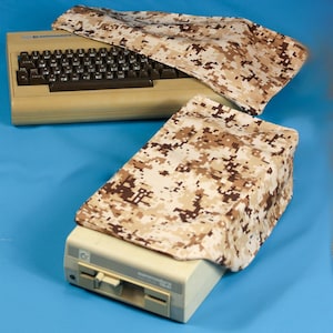 May include: Tan and brown camouflage covers for a vintage Commodore 64 computer and a disk drive. The computer and drive are beige, with black keys and a disk drive slot. The covers are made of a fabric with a pixelated camouflage pattern.