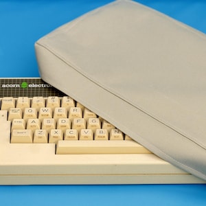 May include: An off-white Acorn Electron computer keyboard with a black label featuring the text acorn electron and a green logo. A rectangular, light gray fabric dust cover is partially draped over the top right section of the keyboard.