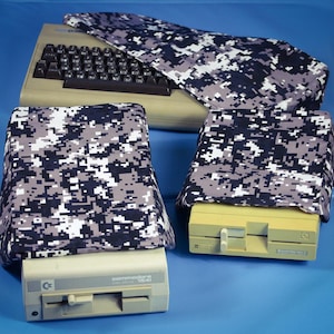 May include: A beige Commodore 64 computer with a black keyboard and two beige Commodore 1541 disk drives. Each device is covered with a digital camouflage fabric cover in shades of gray, black, and white. The background is a solid blue.