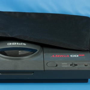Peut inclure: Une console Amiga CD32 grise avec une housse noire partiellement drapée dessus. La console porte les inscriptions "32bit" et "AMIGA CD32" en rouge. Le fond est bleu uni.