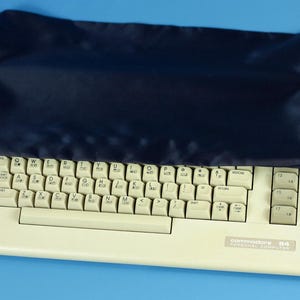 Peut inclure: Clavier d'ordinateur personnel Commodore 64 blanc cassé avec une housse bleu foncé. Le clavier a des touches blanches avec des lettres noires et une étiquette qui dit "Commodore 64 PERSONAL COMPUTER". Le fond est bleu uni.
