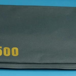 Könnte beinhalten: Ein rechteckiger, dunkelgrauer Stoffkoffer mit dem goldfarbenen Text "A 500". Der Koffer ist gefaltet und scheint aus einem strapazierfähigen Material zu bestehen. Der Hintergrund ist einfarbig blau.