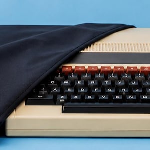 May include: A beige BBC Master 128 computer with a black keyboard, a row of red function keys, and a black fabric cover draped over the left side. The computer features a black horizontal panel above the keys with the text BBC Master 128.
