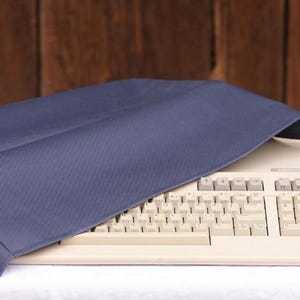 May include: A beige computer keyboard partially covered by a dark blue fabric dust cover. The keyboard features a standard QWERTY layout with function keys along the top and a numeric keypad on the right side, with visible text on the keys.