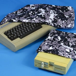 May include: Two beige Commodore 64 computers with black keyboards and a matching digital camouflage cover. The covers are black, white, and gray. The computers are on a blue surface.