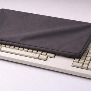 Puede incluir: Un teclado de ordenador Atari vintage con teclas blanquecinas y una funda gris oscuro. El logotipo de Atari es azul. Una pieza clásica de tecnología.